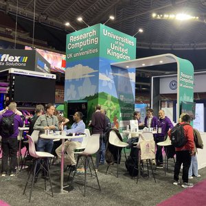 People standing and sitting at a green and blue coloured exhibition booth which has some AV screens in it. Everyone is talking to someone.