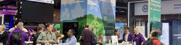 People standing and sitting at a green and blue coloured exhibition booth which has some AV screens in it. Everyone is talking to someone.