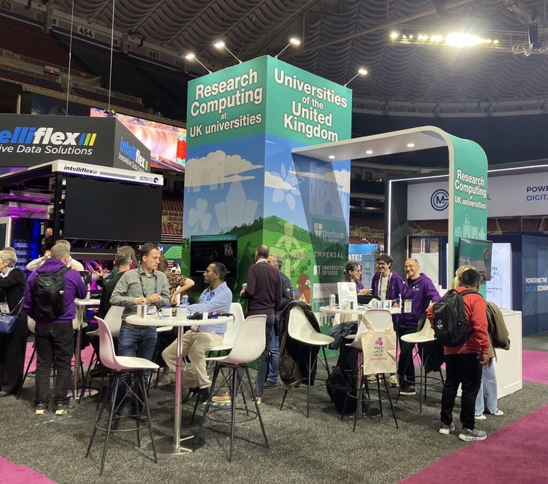 People standing and sitting at a green and blue coloured exhibition booth which has some AV screens in it. Everyone is talking to someone.