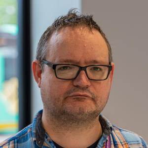 Head and shoulders image of Andrew.  He has stubble and wears glasses.  He's wearing a light blue tartan shirt.