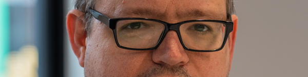 Head and shoulders image of Andrew.  He has stubble and wears glasses.  He's wearing a light blue tartan shirt.