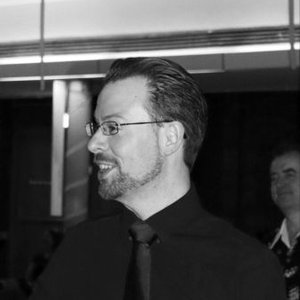 Head and shoulders photo in black and white of Ben.  He's turned slightly to the left hand side.  He has short dark hair and a short but full beard.  He is wearing dark rimmed glasses and a dark coloured long sleeve shirt.