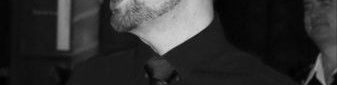 Head and shoulders photo in black and white of Ben.  He's turned slightly to the left hand side.  He has short dark hair and a short but full beard.  He is wearing dark rimmed glasses and a dark coloured long sleeve shirt.