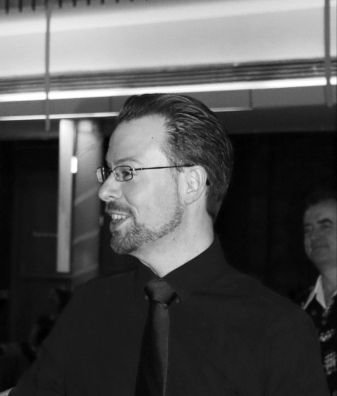 Head and shoulders photo in black and white of Ben.  He's turned slightly to the left hand side.  He has short dark hair and a short but full beard.  He is wearing dark rimmed glasses and a dark coloured long sleeve shirt.