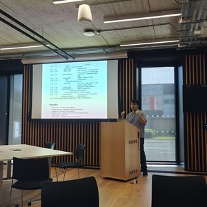 A male speaker standing in front of a projection screen at a lectern talking to a room where people are sitting round tables facing him