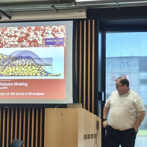 A male speaker standing in front of a projection screen at a lectern talking to a room where people are sitting round tables facing him