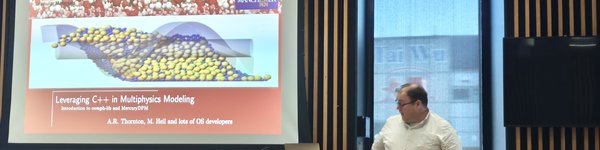 A male speaker standing in front of a projection screen at a lectern talking to a room where people are sitting round tables facing him