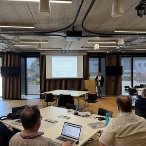 A male speaker standing in front of a projection screen at a lectern talking to a room where people are sitting round tables facing him