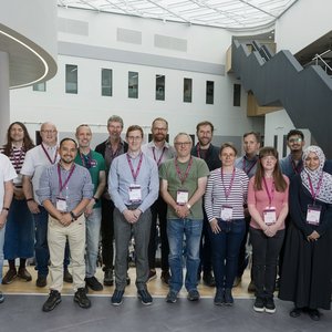 A group of 17 people from UoM at a conference.  They are standing facing the camera wearing conference badges and a wide variety of clothing!.