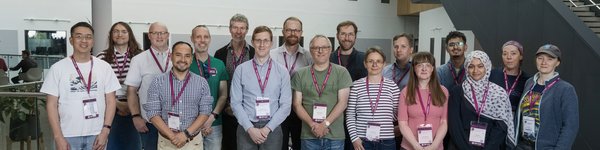 A group of 17 people from UoM at a conference.  They are standing facing the camera wearing conference badges and a wide variety of clothing!.