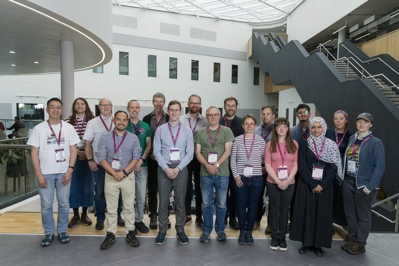 A group of 17 people from UoM at a conference.  They are standing facing the camera wearing conference badges and a wide variety of clothing!.