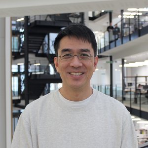 A head and shoulders photo of Raymond. He is Japanese and has dark short hair and is wearing black framed glasses and a grey jumper.  He is smiling.