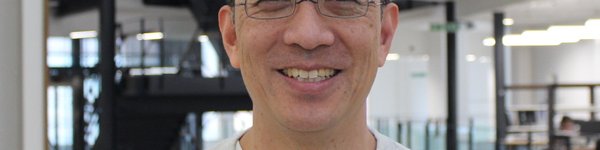 A head and shoulders photo of Raymond. He is Japanese and has dark short hair and is wearing black framed glasses and a grey jumper.  He is smiling.