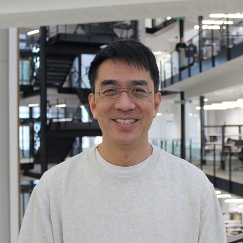 A head and shoulders photo of Raymond. He is Japanese and has dark short hair and clear framed glasses.  He is wearing a light grey jumper with a white t-shirt and he is smiling. The background is a foyer of an open plan building with stairs