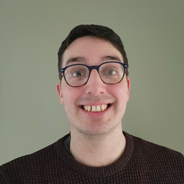 Head and shoulders photo of a white man with short black hair.  He is wearing black rimmed glasses and is grinning widely.  He is wearing a black crew neck jumper.
