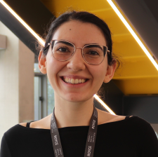 Head and shoulders photo of Stavrina - a woman with dark hair in a ponytail. She is smiling broadly and is wearing gold thin framed glasses and a black boat-necked top.