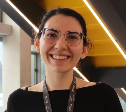 Head and shoulders photo of Stavrina - a woman with dark hair in a ponytail. She is smiling broadly and is wearing gold thin framed glasses and a black boat-necked top.