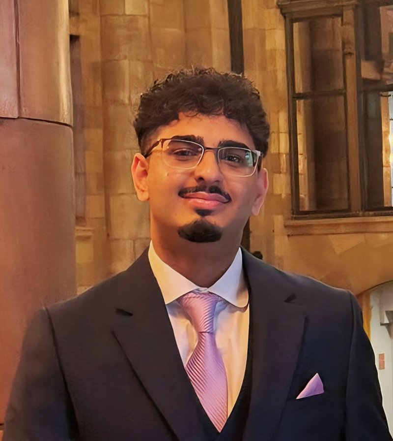 Head and shoulders photo of a young man. He has short black slightly curly hair, silver rimmed glasses and has a thin black moustache and a small black goatee beard.  He is wearing a dark coloured suit, white shirt and pale lavender coloured tie.