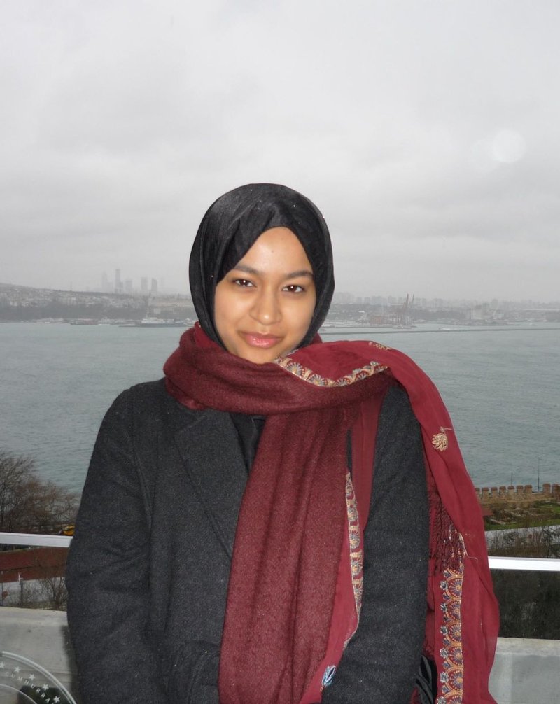 A photo of a young woman standing on a balcony with a sea port in the distance.  It is a grey and hazy day.  She is wearing a dark blue hijab with a dark blue suit jacket.  She also has a dark red scarf wrapped round her neck.