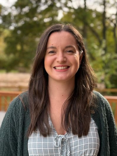 Head and shoulder photo of Sian who is a woman with long dark brown hair and is smiling. She is wearing a dark green cable knit cardigan and a blue and white checked top that ties at the front. The background is slightly out of focus trees.