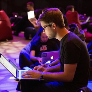 A young man sits in the dark on a chair typing on a laptop