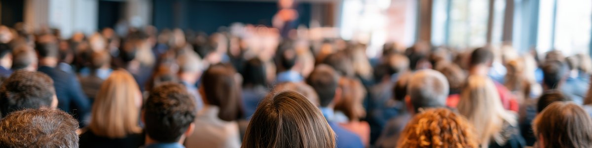 A group of people at a conference.  They are sitting down and the photo was taken from behind so you just see the back of their heads. They are all looking at projection screen.