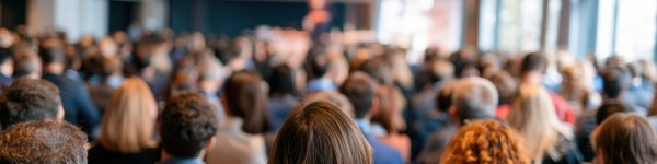 A group of people at a conference.  They are sitting down and the photo was taken from behind so you just see the back of their heads. They are all looking at projection screen.
