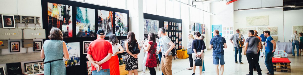 People in bright white building milling around looking at paintings hung on black boards