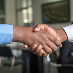 The hands and arms of 2 men shaking hands.  Both men are black with one man wearing a white shirt and the other man wearing a blue shirt.