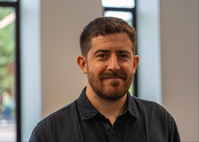 A head and shoulders photo of Martin. A male with short brown hair and a short full beard wearing a collared shirt standing in a white walled office with vertical windows.