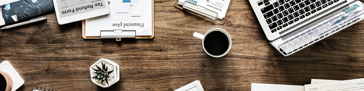 A brown wooden desk seen from above.  There is an open silver laptop on it, a cup of coffee, and pages of white A4 paper with planning details and writing on them scattered about the desk.