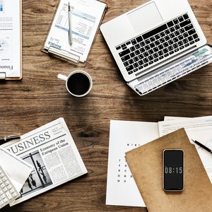 A brown wooden desk seen from above.  There is an open silver laptop on it, a cup of coffee, and pages of white A4 paper with planning details and writing on them scattered about the desk.